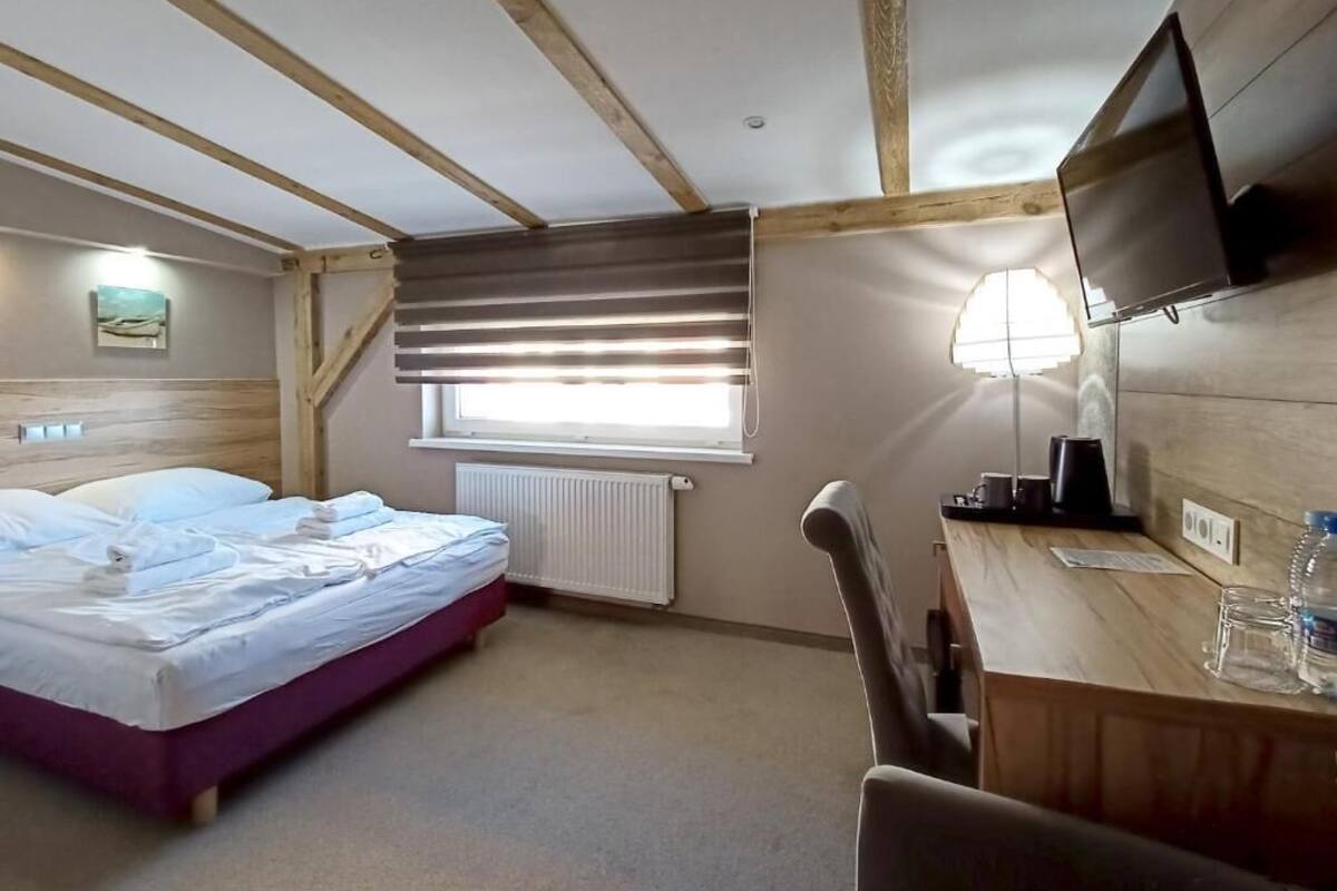 Une chambre à coucher comprenant un lit, un bureau avec une chaise, un téléviseur, une fenêtre avec des stores et un radiateur.