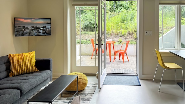 Ein modernes Wohnzimmer mit grauem Sofa, gelbem Kissen und einem schwarzen Couchtisch. Ein großes Fenster öffnet sich zu einer Terrasse im Freien mit orangefarbenen und gelben Stühlen.