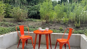 Ein kleiner Sitzbereich im Freien mit zwei orangefarbenen Stühlen und einem Tisch, umgeben von Pflanzen.