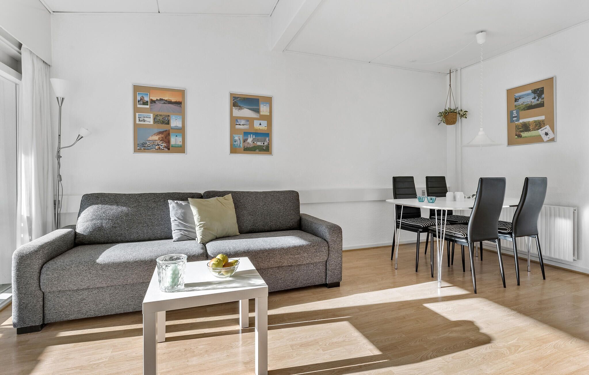 A modern living room with a grey sofa, a small white table with a glass and fruit bowl, and a dining area with a white table and black chairs. Two framed pictures adorn the walls.