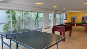 A room with a ping pong table, foosball table, and pool table, large windows with a view of palm trees, and a yellow wall with artwork. - Bela Hospedagem - Jd do Atlântico 402B (João Pessoa)