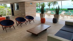A modern lounge area with wicker furniture, a wooden coffee table, and a view of the ocean. - Bela Hospedagem - Jd do Atlântico 402B (João Pessoa)