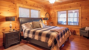 Un dormitorio de cabaña de madera con cama, mesitas de noche, una silla y un ventilador de techo.