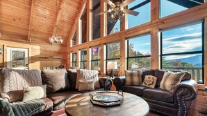 A cozy cabin interior with wooden walls, large windows, a leather sofa, and a round table.