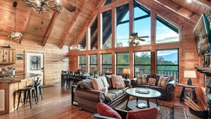 A spacious log cabin interior with a large living area, high ceilings, and large windows offering views of the outdoors.
