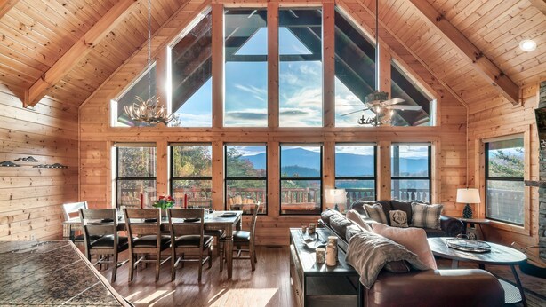 A spacious wooden cabin with a high ceiling, large windows offering mountain views, a dining area, and a cozy living room with a fireplace.