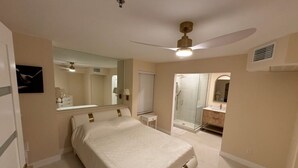 Ein Schlafzimmer mit Bett, Deckenventilator, Badezimmer mit Dusche und Glastür zum Balkon.