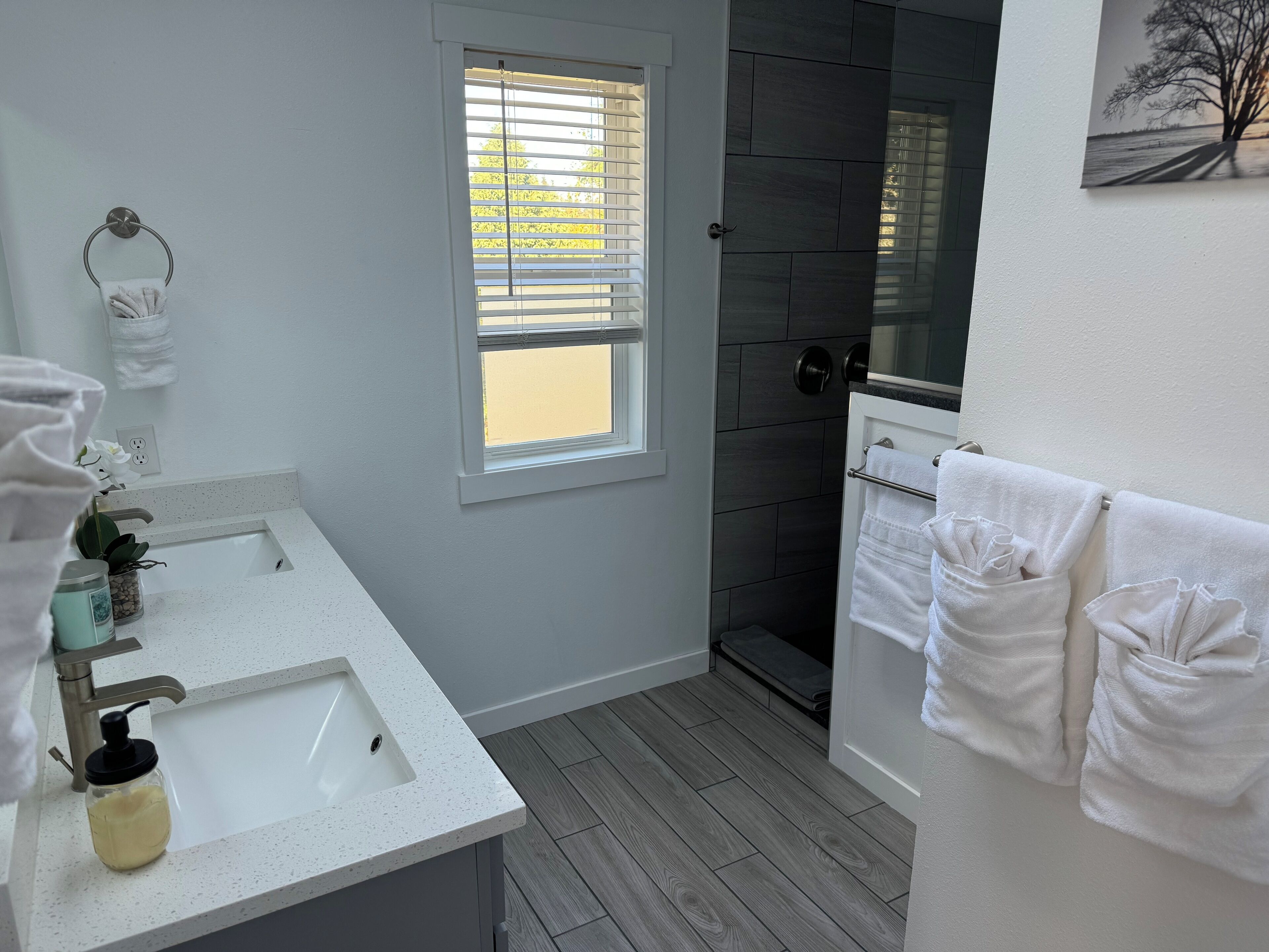 A modern bathroom with a white sink, a window with blinds, a shower area, and towels hanging on a rack.