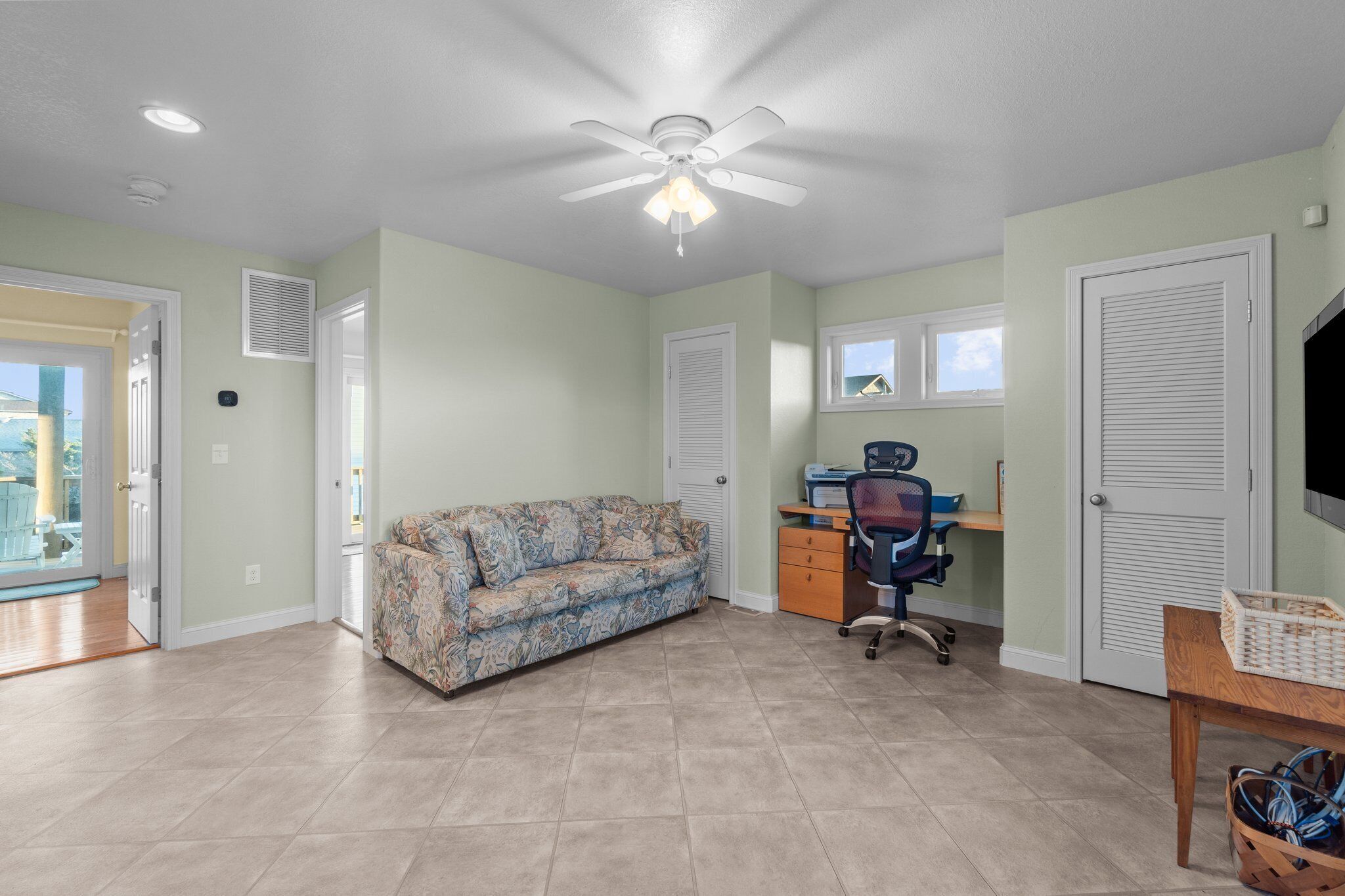 A well-lit room with a ceiling fan, a desk with a chair, a patterned sofa, and a TV on the wall.
