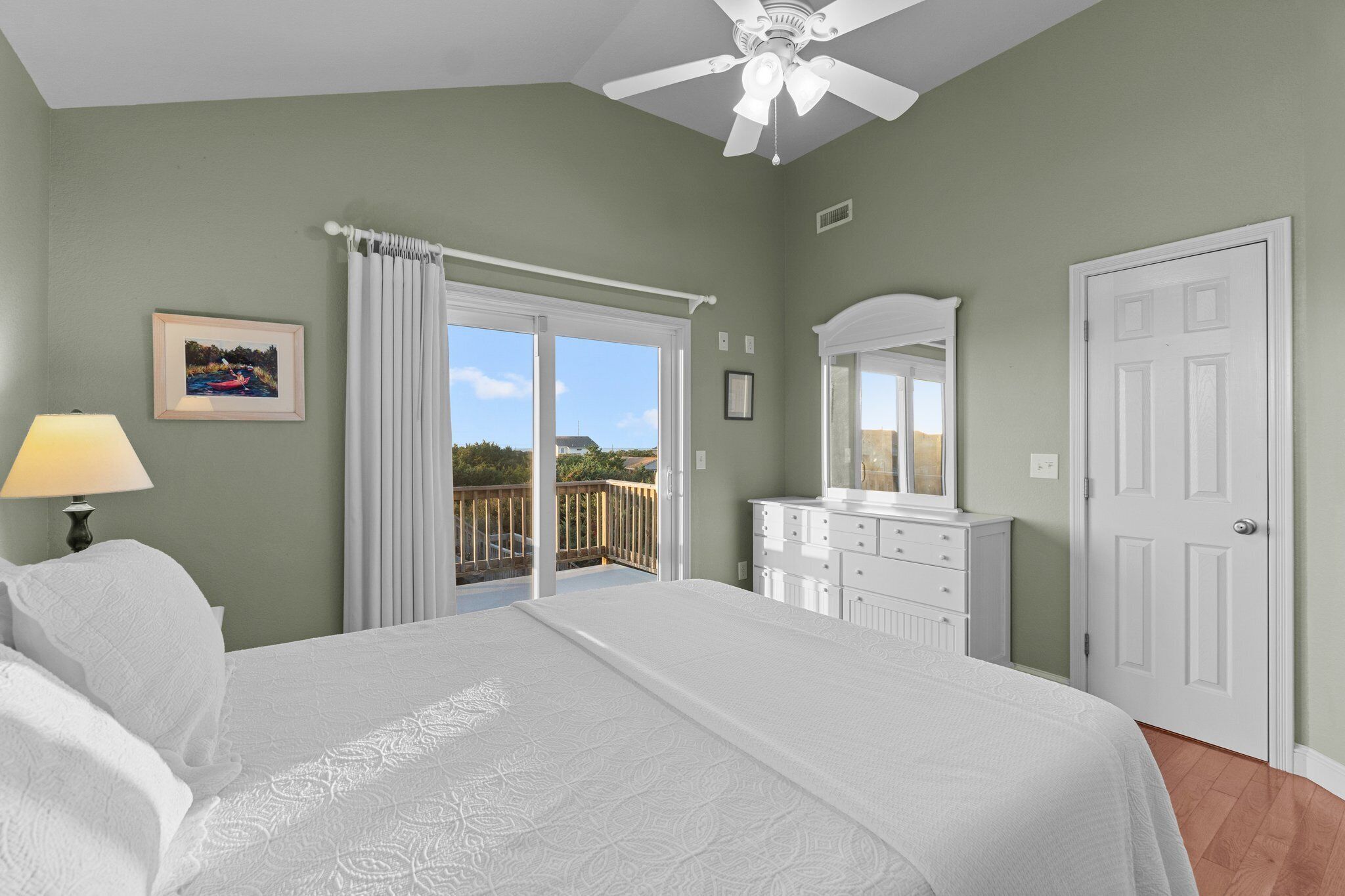 A bedroom with a bed, a dresser, a ceiling fan, and a balcony with a view.