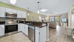 A modern kitchen with white cabinets, a large island, stainless steel appliances, and a dining area with a sofa and TV.