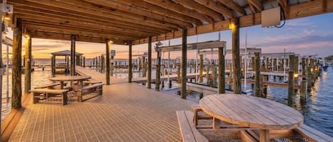 Terraza de madera con mesas de picnic y bancos que da vista a un puerto deportivo.