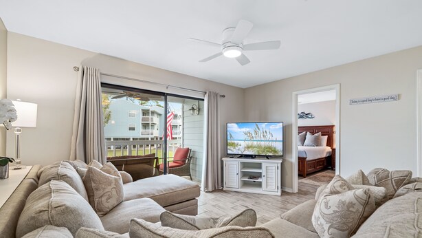 A living room with a large sectional sofa, a flat-screen TV, and a ceiling fan. There is a sliding glass door leading to a balcony with a view of a building and a flag.