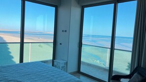 A hotel room with a large bed, a chair, and a view of the beach through large windows.