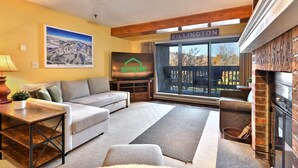 A cozy living room with a sofa, a coffee table, a fireplace, and a large window overlooking a deck.