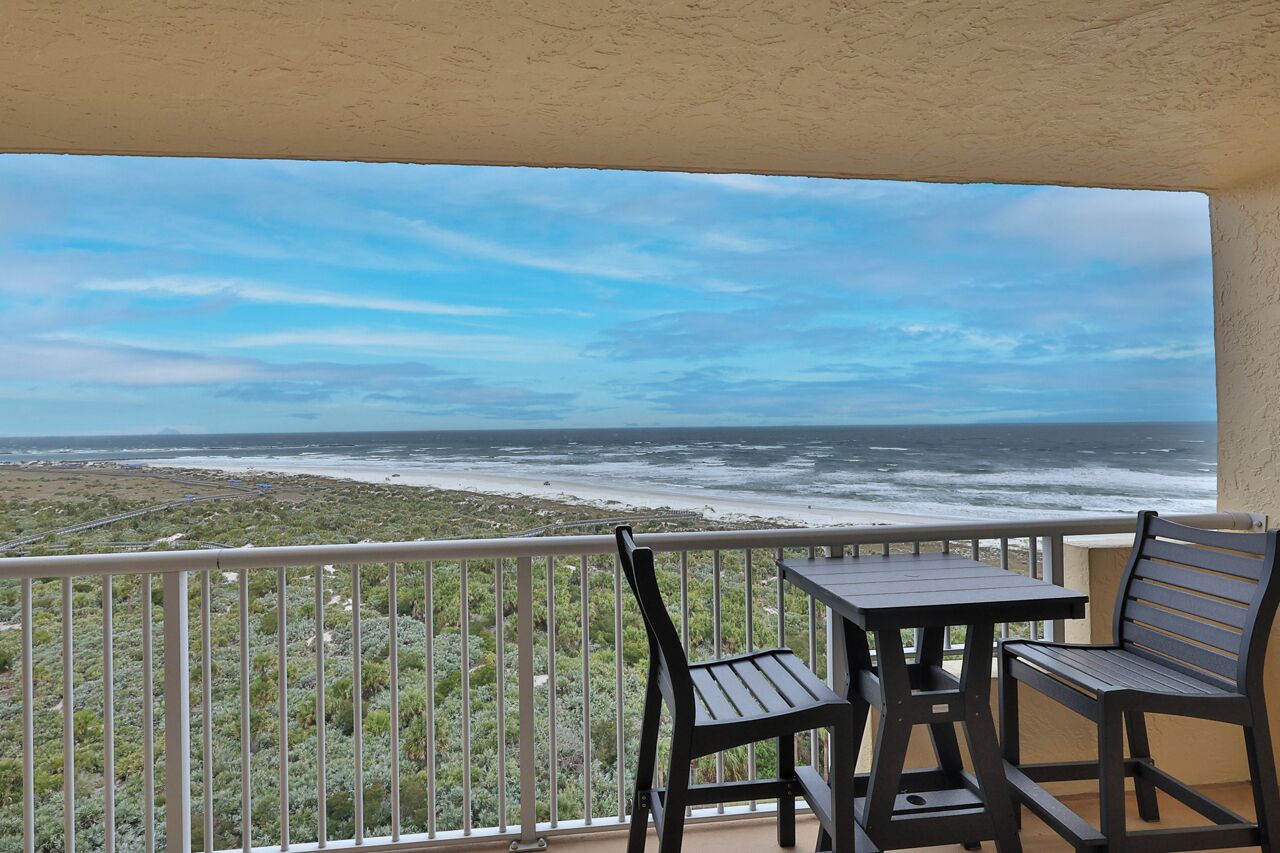 A balcony with a table and two chairs overlooking a beach and ocean.