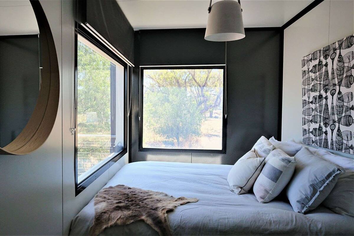 A modern bedroom with a large bed, two windows, a circular mirror, and a wall art piece.