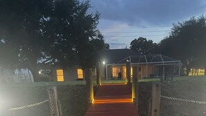 Una terraza de madera con escalones que conducen a un área cubierta, rodeada de árboles y un edificio al fondo.