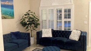 Una sala de estar con dos sofás azules, un otoman redondo, un ventilador de techo, un ventanal con persianas, una planta en maceta y una alfombra estampada.