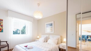 A bedroom with a large bed, bedside tables, a window with blinds, a hanging light fixture, and a view of a blue sofa and chair through an open door. - A-Br – Quiet, Infinity Pool & Private Parking (Cannes)