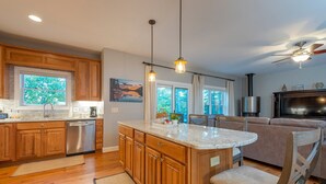 Una cocina moderna con armarios de madera, un lavavajillas de acero inoxidable y una isla grande. Hay un comedor con un sofá y una silla.