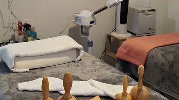 Una stanza per massaggi con un lettino da massaggio, strumenti di legno per massaggi, un asciugamano bianco, una lampada per massaggi e uno scaffale con flaconi e un vaso.