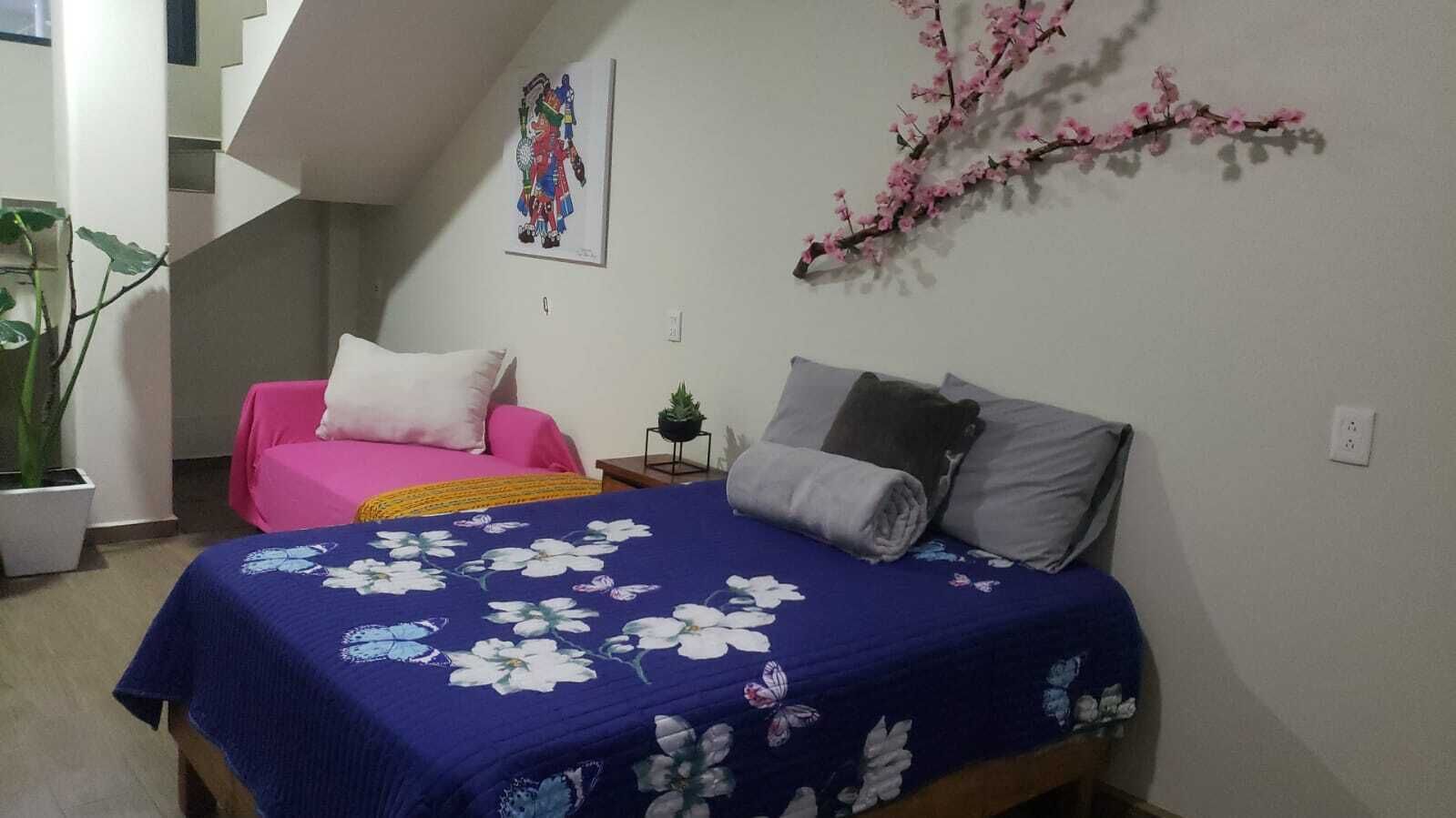 Ein Schlafzimmer mit einem Bett, einem rosa Sessel, einem kleinen Tisch mit einer Pflanze und einer Treppe im Hintergrund.