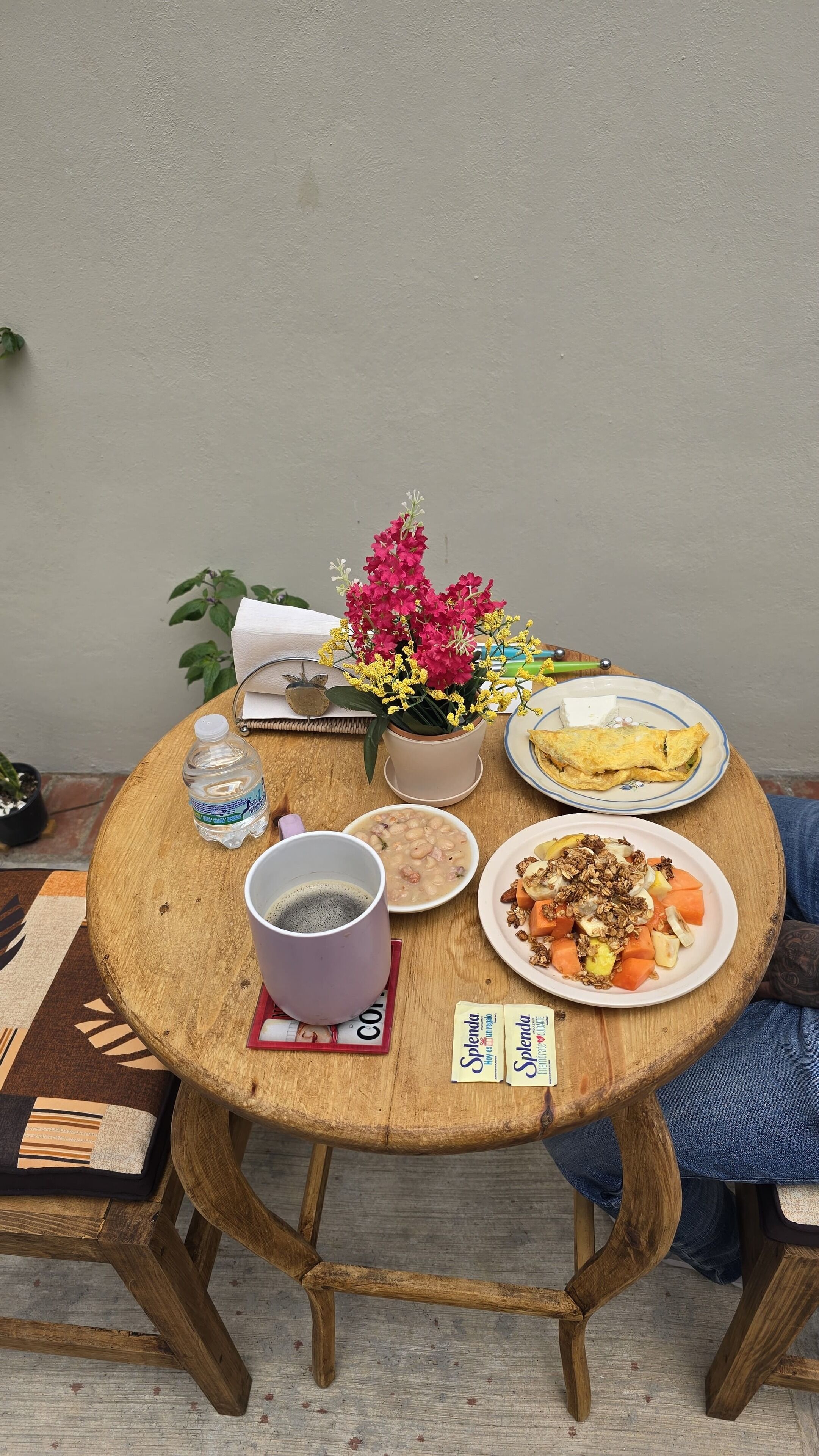 Een ronde houten tafel met diverse soorten eten, een potplant met roze bloemen, een waterfles en een kop koffie.