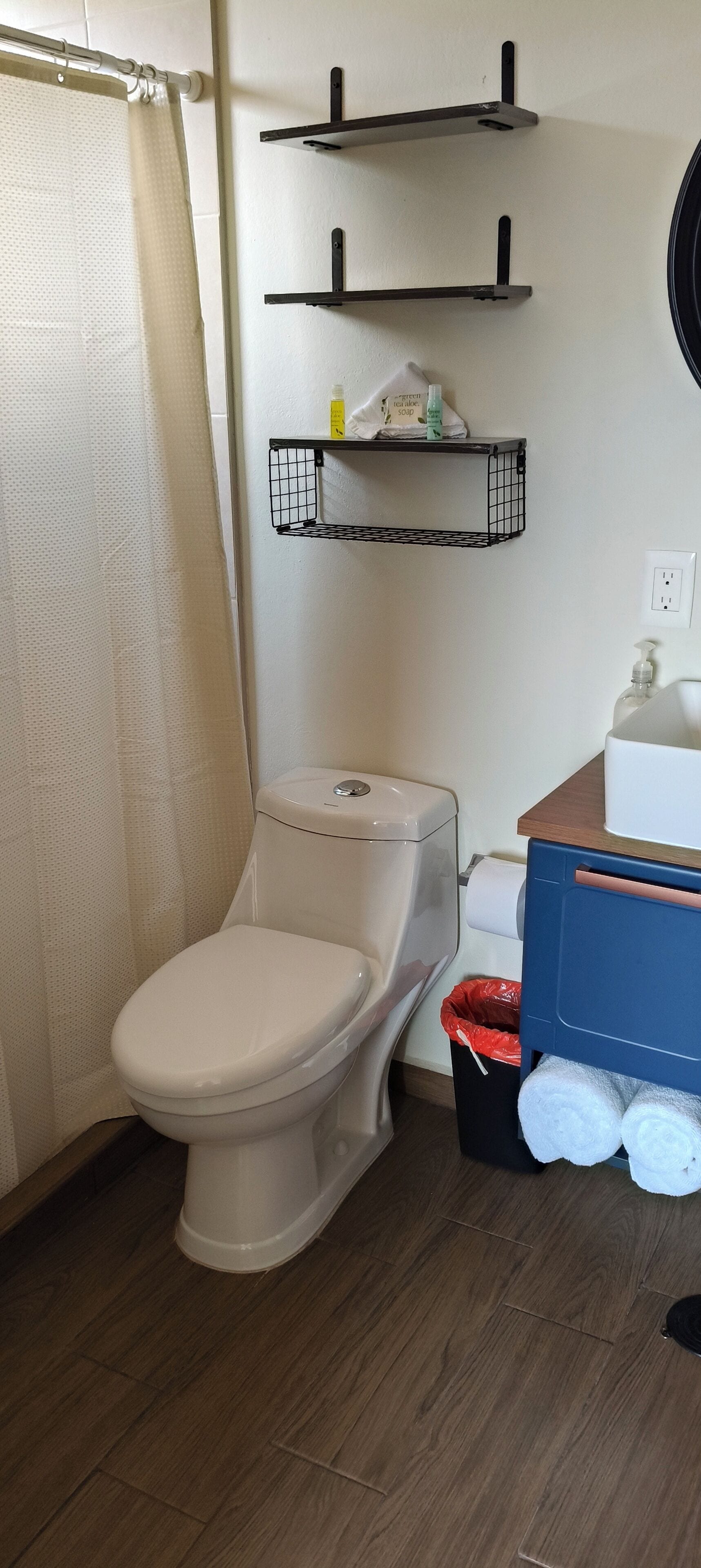 Ein Badezimmer mit Toilette, Duschvorhang, Mülleimer und Ablagen mit Pflegeprodukten.