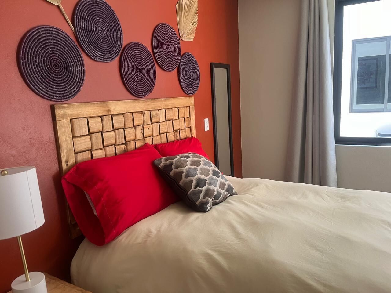 Una cama con cabecera de madera, almohadas rojas con estampado, una lámpara blanca y decoración en la pared.