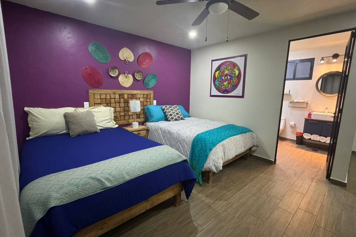 A bedroom with two beds, a wooden headboard, a ceiling fan, and a bathroom visible through an open door.