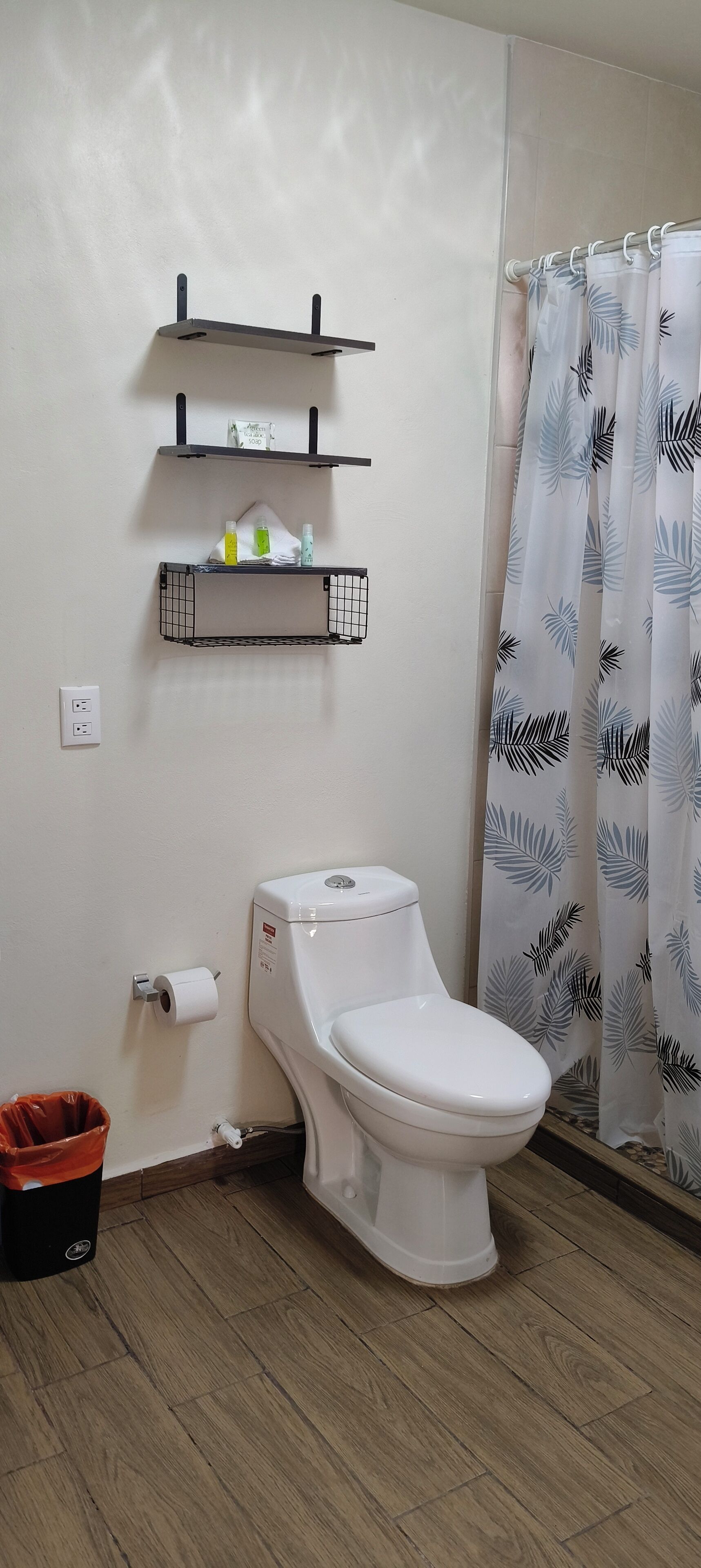 A bathroom with a toilet, a shelf with toiletries, a trash bin, and a shower curtain.