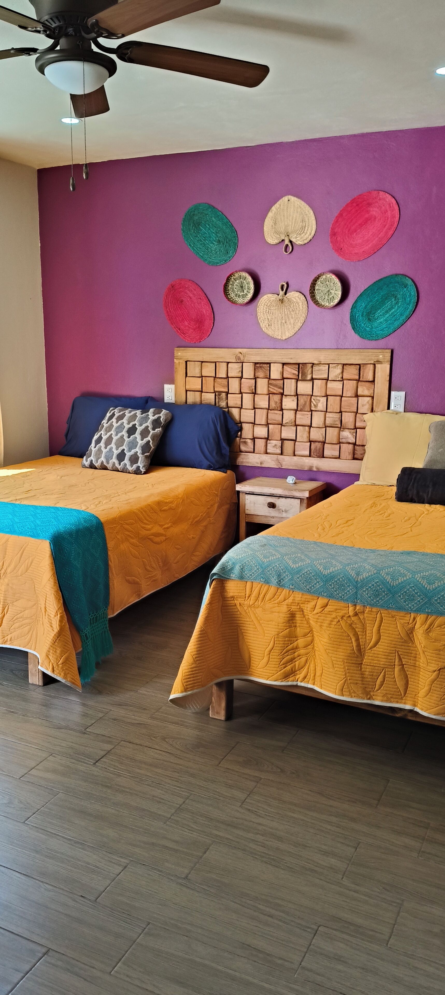 Quarto com duas camas, cabeceira de madeira, ventilador de teto e parede decorada com padrões circulares em tecido.