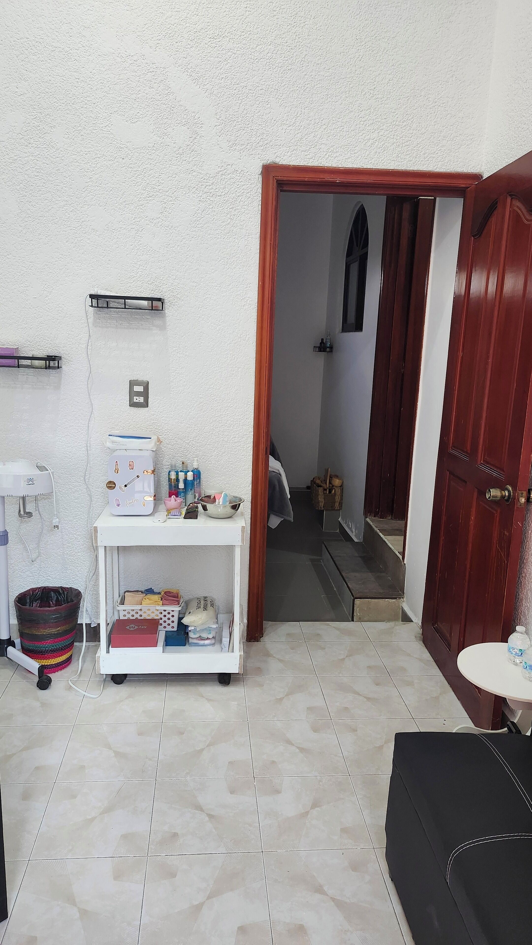 Una habitación pequeña y limpia, con piso de baldosas blancas, un estante blanco con artículos de aseo, una puerta de madera y una mesita con una botella.