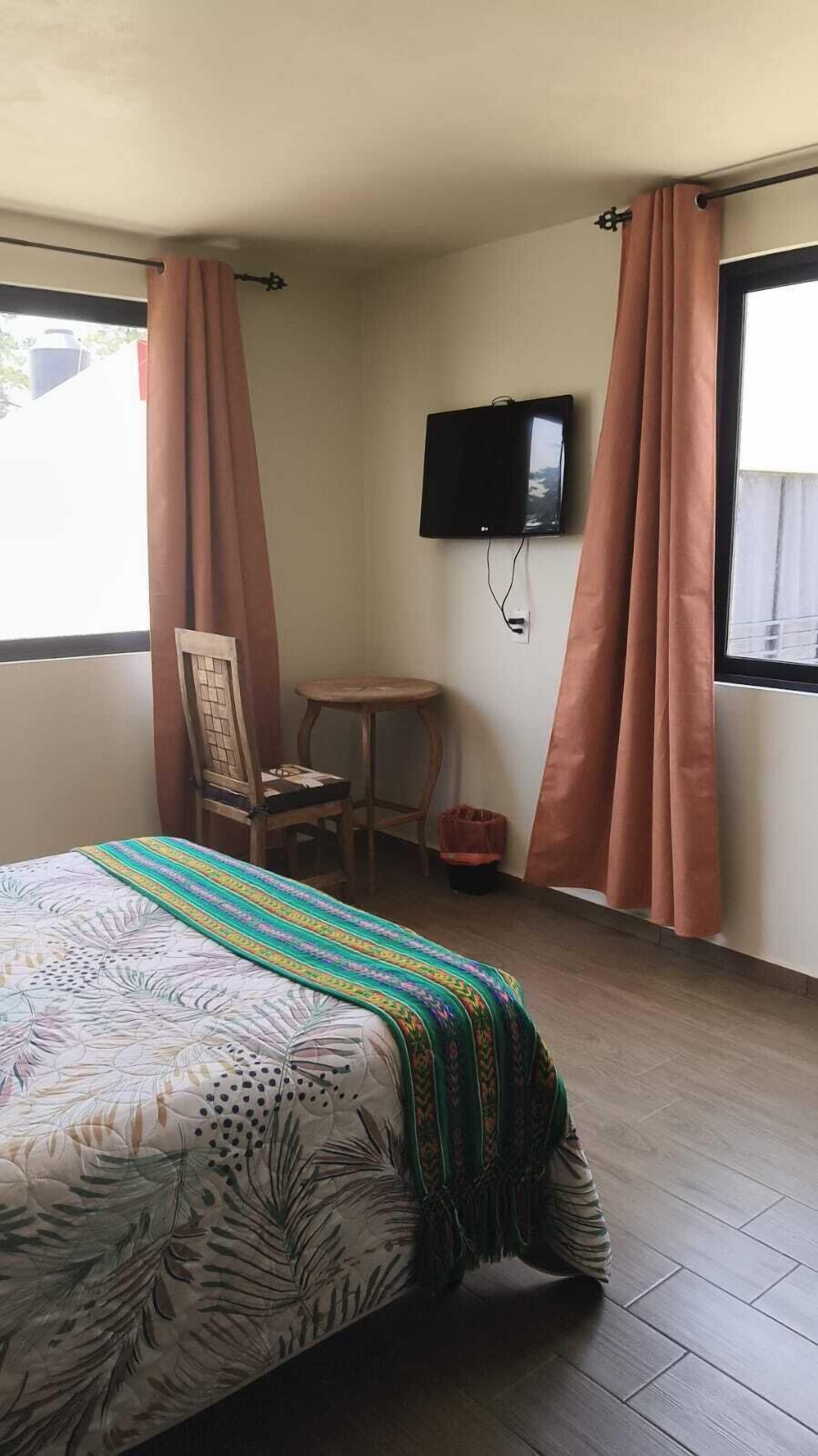 Una habitación con una cama, televisión montada en la pared, una silla y una mesita.