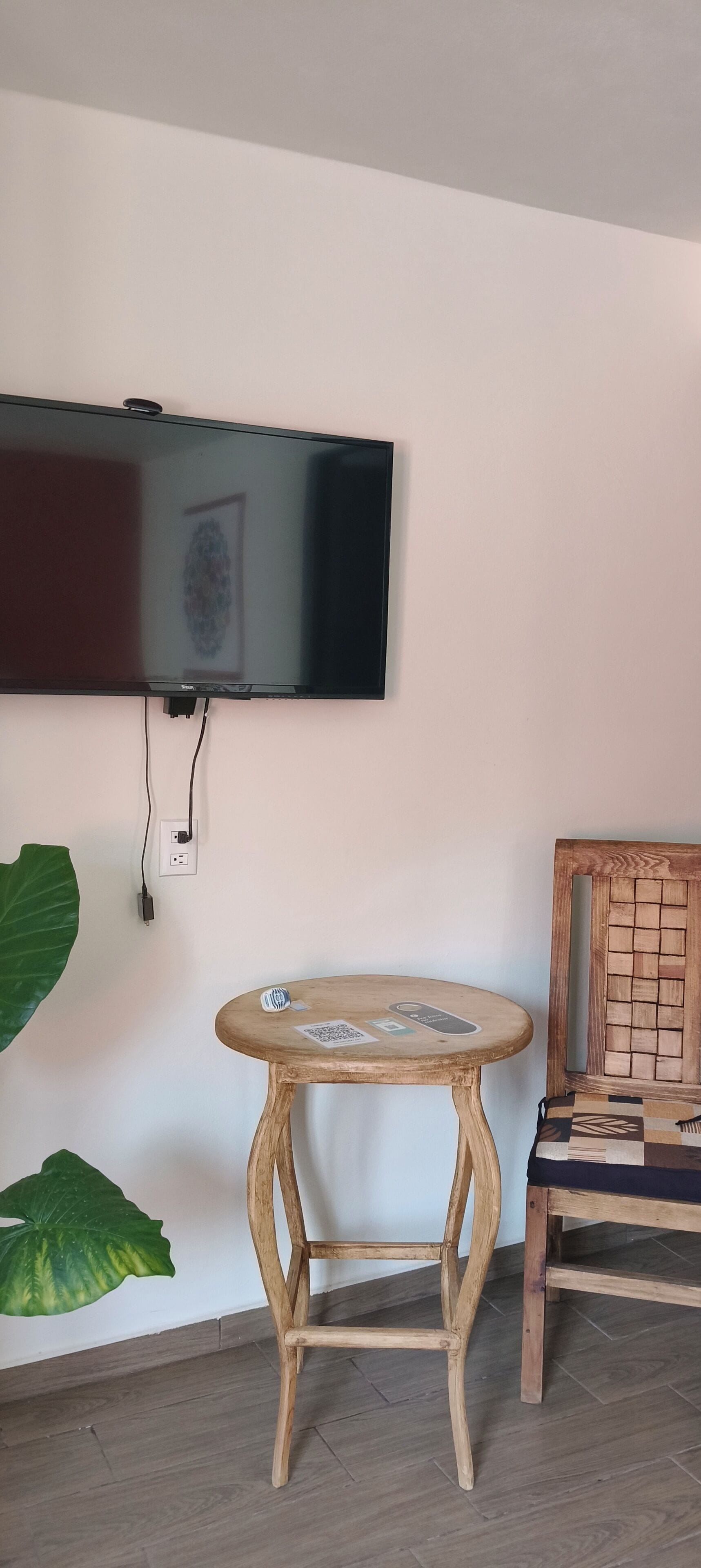 A room with a flat-screen TV mounted on the wall, a round wooden table with a QR code, and a wooden chair with a patterned cushion.