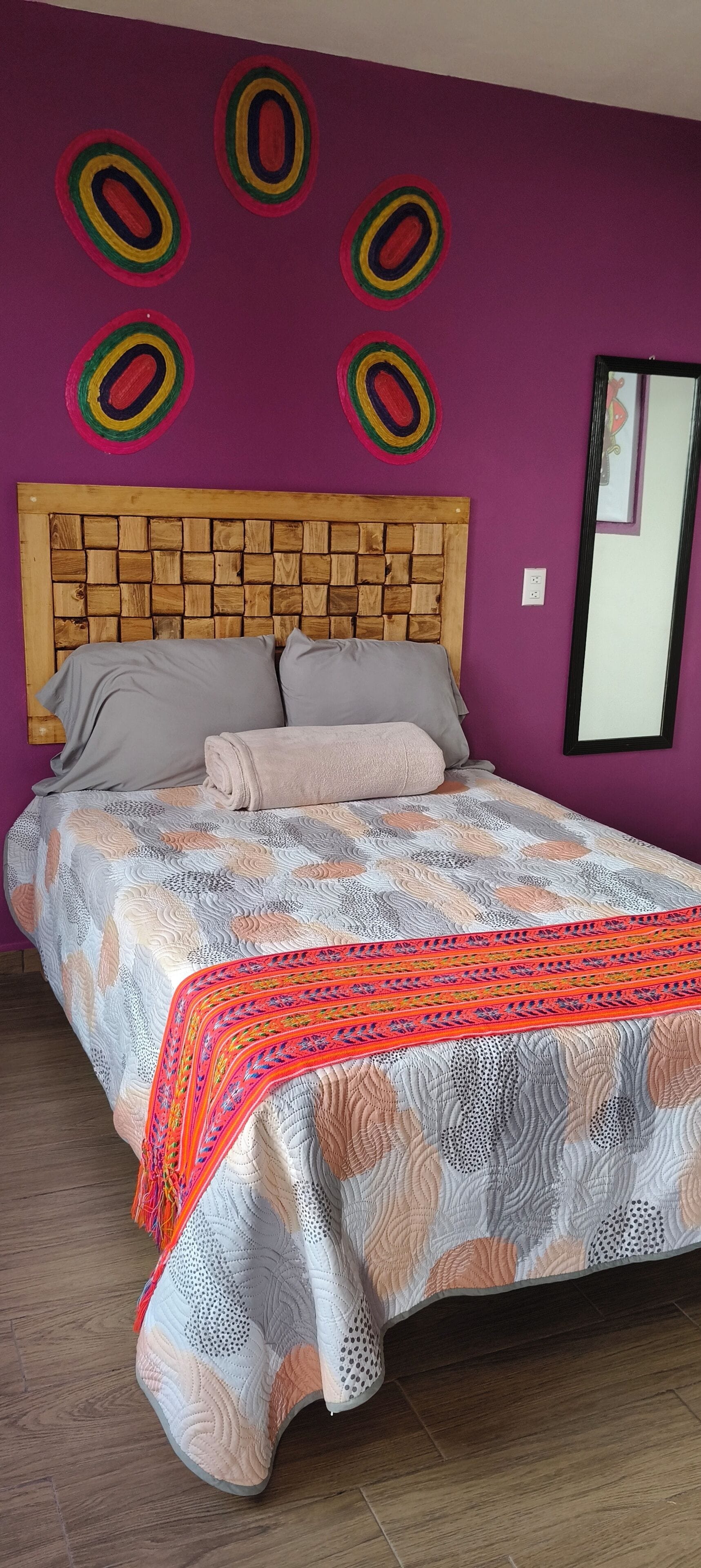 Una cama con colcha estampada y almohadas, cabecera de madera y un espejo en la pared.