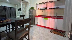 Une cuisine compacte avec un plan de travail rouge, des armoires blanches et une cuisinière intégrée. On y trouve un coin repas avec une table et des chaises, ainsi qu’un escalier en colimaçon menant à un étage supérieur.
