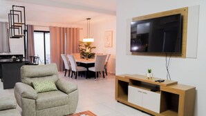 A modern living room with a sofa, a coffee table, a TV mounted on the wall, a dining area with a table and chairs, and a kitchen area in the background. - Beautiful Modern Home in Res San Andres  (San Miguel)