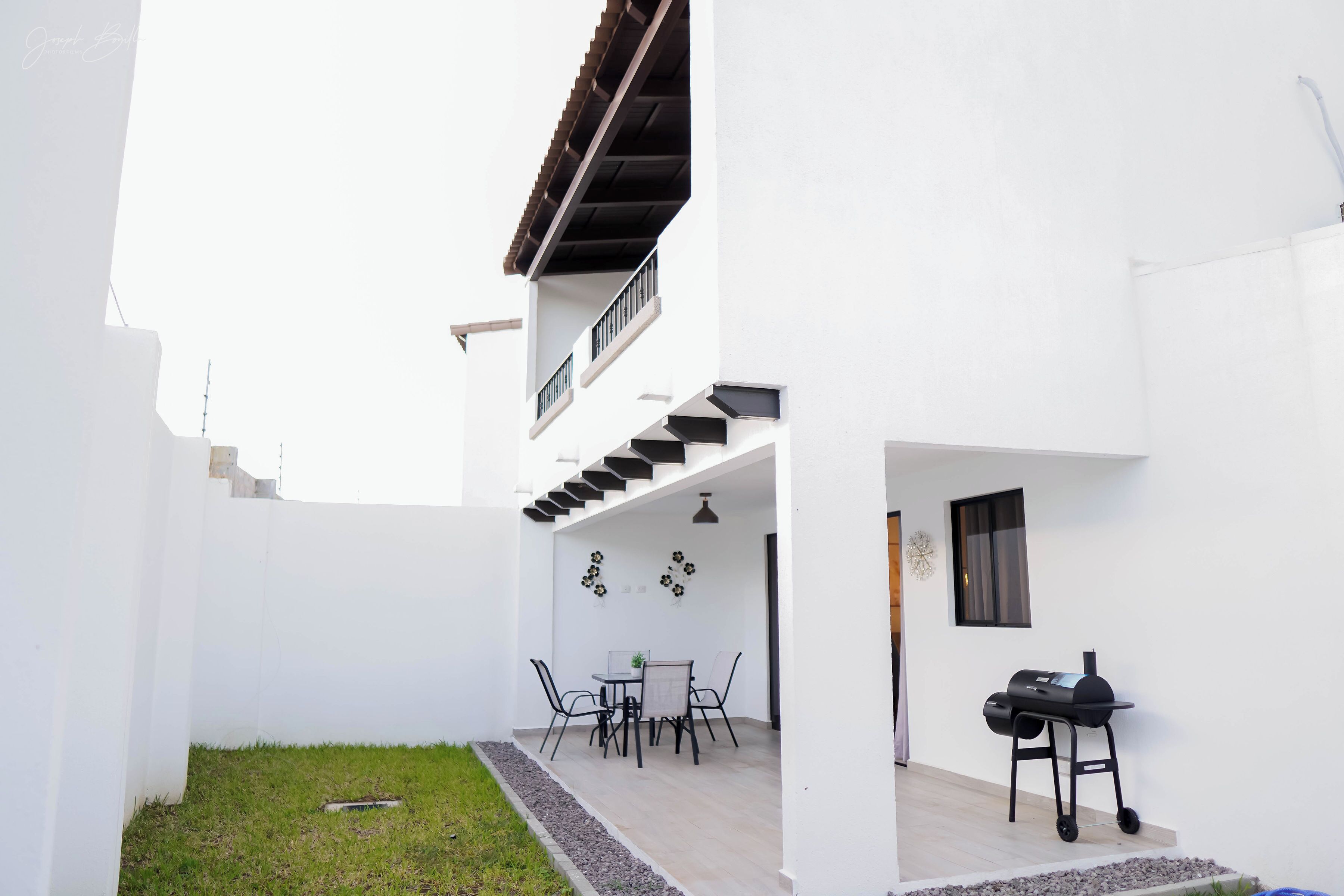 Un edificio blanco con un patio que tiene una mesa, sillas, una parrilla y un pequeño jardín.
