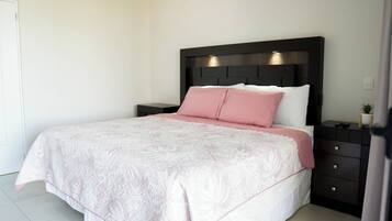 Un lit bien fait, avec une couvre-lit rose et blanche, une tête de lit sombre et une petite table de chevet agrémentée d’une plante.