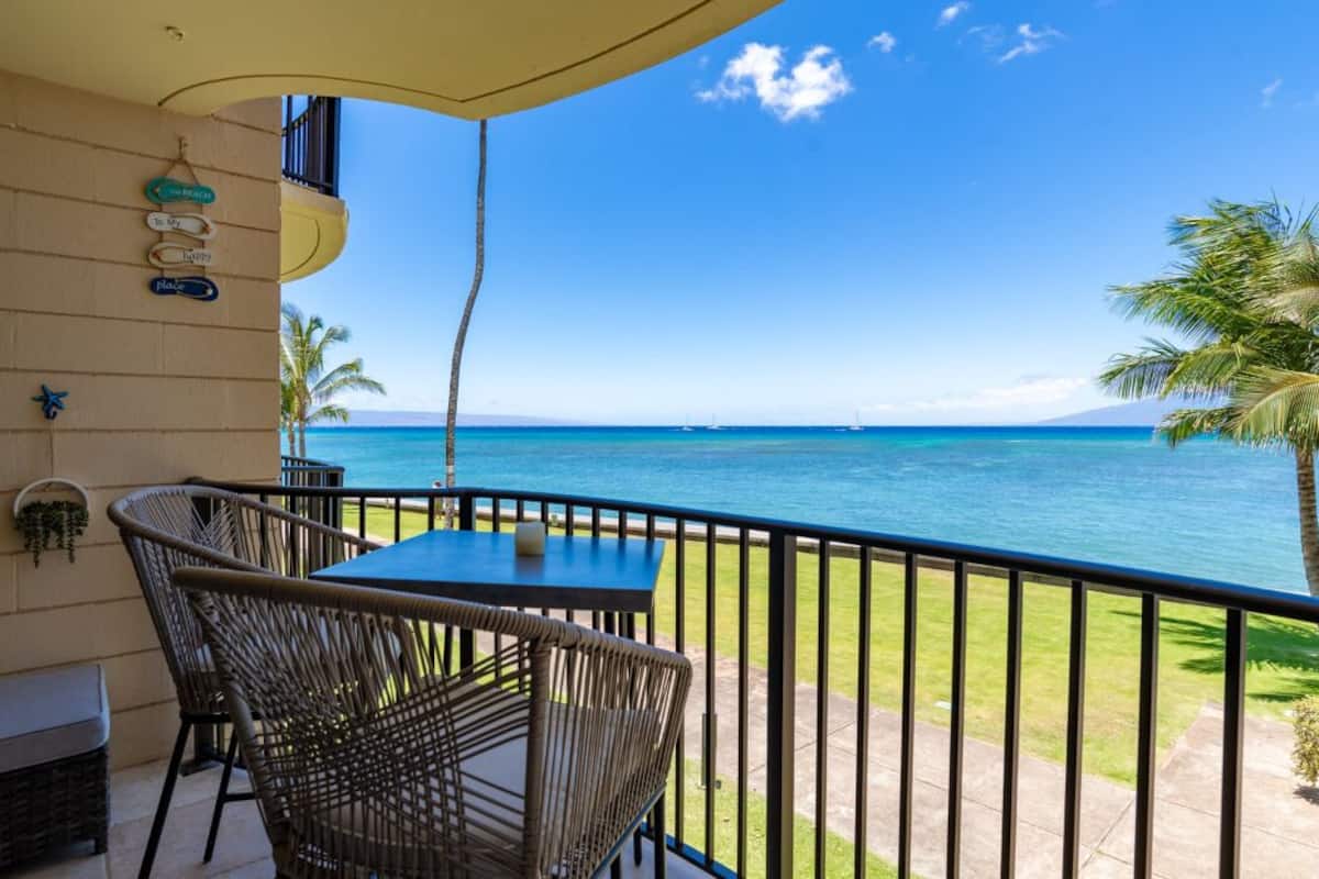 Un balcon agrémenté de fauteuils et d’une table en osier, d’où l’on peut admirer une mer bleue et limpide, ainsi que des bateaux et une montagne lointaine.
