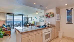 Una cocina moderna con una isla grande, microondas y lavavajillas. Hay una zona de comedor con sillas y una mesa, y vistas al océano a través de ventanas amplias.
