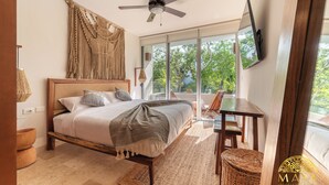 A bedroom with a bed, a ceiling fan, a large window, and a view of greenery outside. - NHOA 209B Lux 1BR Pool View Aldea Zama (Tulum)