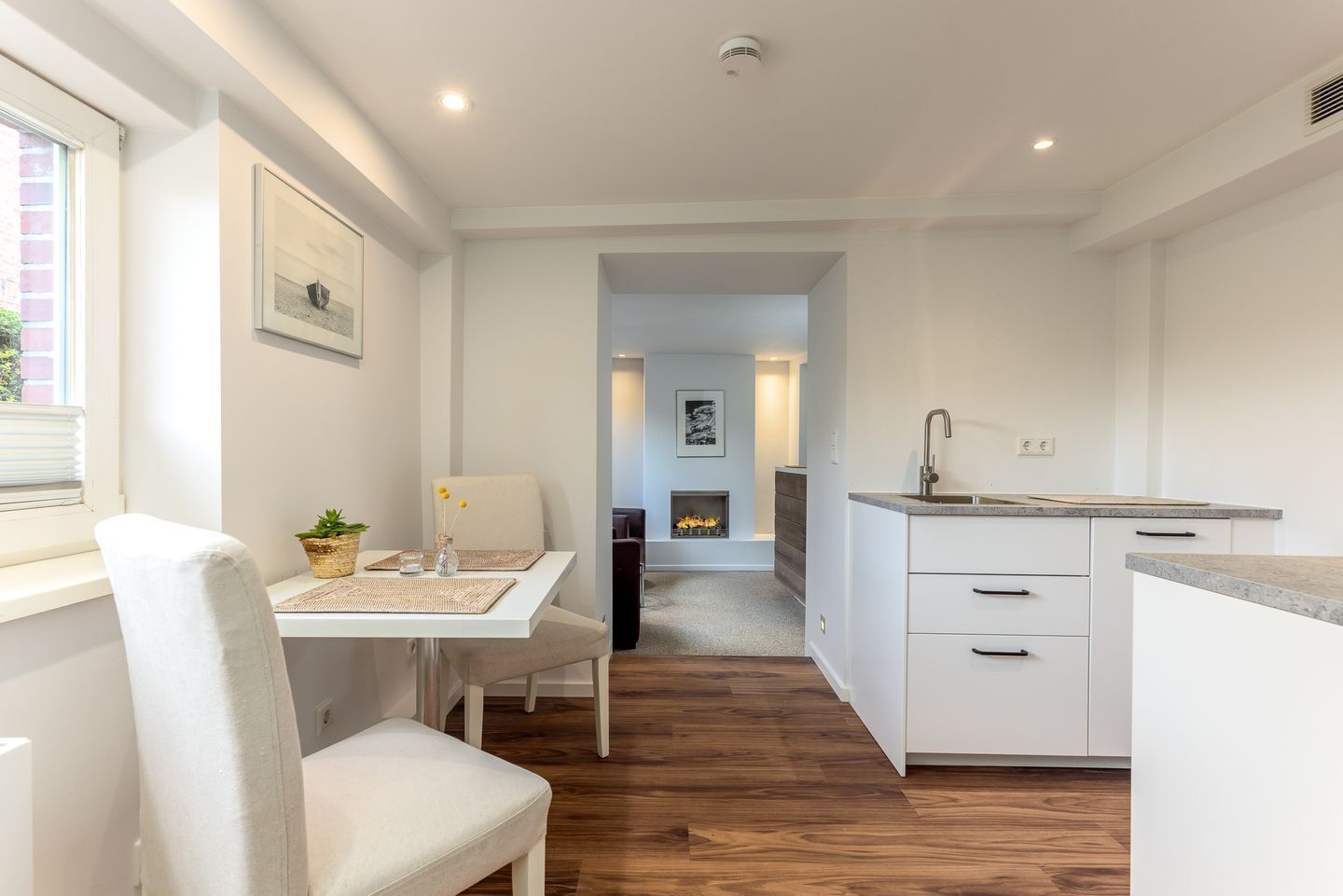 A modern kitchen with white cabinetry, a dining area with a white table and chair, and a fireplace in the background.