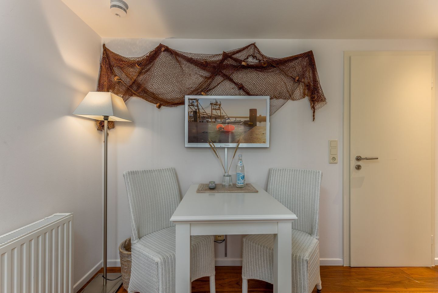 A small dining area with a white table and two wicker chairs, a framed picture above the table, and a floor lamp to the left.