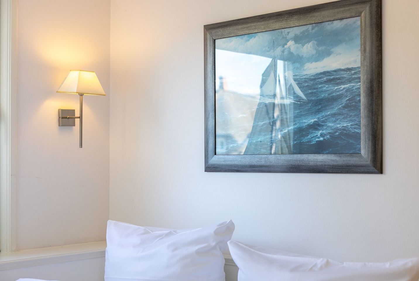 Um quadro emoldurado com um veleiro no mar, um abajur fixado na parede e uma cama branca com almofadas.