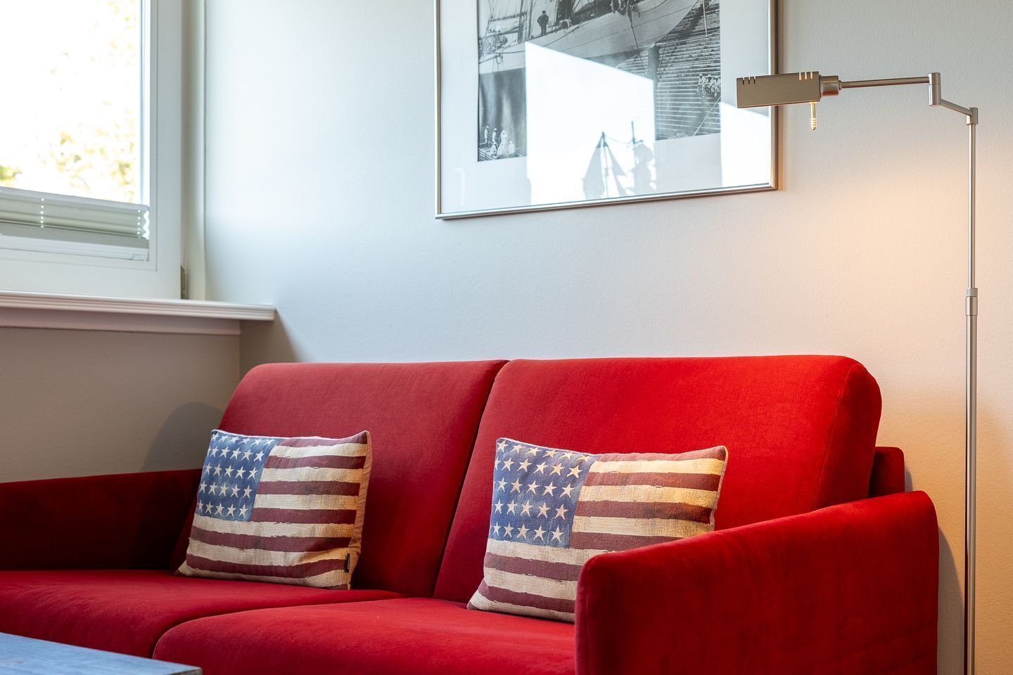A red sofa with two pillows featuring the American flag design, a floor lamp, and a framed picture on the wall.