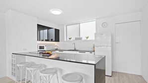 Una cocina moderna con una isla central, taburetes blancos de bar, un refrigerador y una ventana con un reloj.