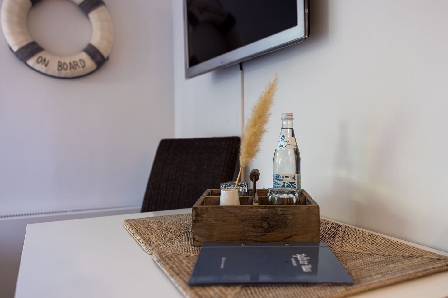 Una caja de madera con una botella, un vaso y una vela sobre una mesa, con un televisor y un salvavidas en la pared.